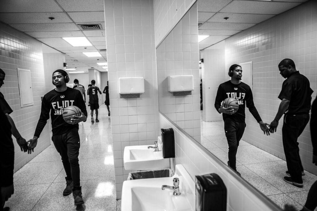 Black and white photo of a young basketball player holding a basketball and wearing a 'Flint Jaguars' shirt, walking through a tiled hallway while wearing headphones. He exchanges a handshake with a man as he walks past. Other players can be seen in the background, some reflected in a mirror on the wall.