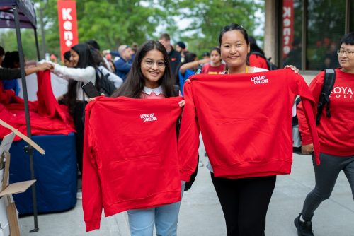 Two students posing with red Loyalist hoodies. In the background, more students stand in line, waiting for red hoodies.