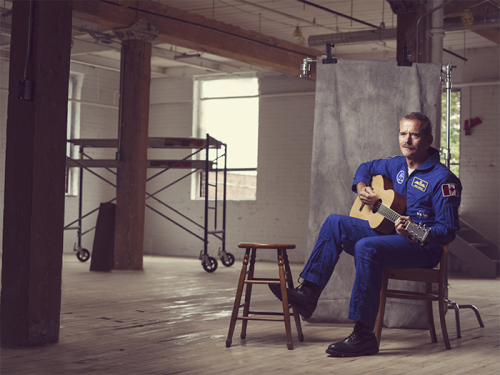 Colonel Chris Hadfield in an empty old building, posed on a chair in his flight suit with a guitar on his lap.