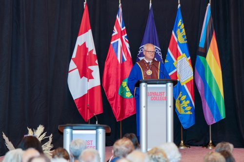 Mark Kirkpatrick in ceremonial robes, standing at a Loyalist College podium in front of a black backdrop and several flags.