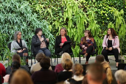 A group of women are sitting on chairs on a raised platform, one is holding a microphone and talking. They're all sitting in front of a live plant green wall in Loyalist's Link Lounge.