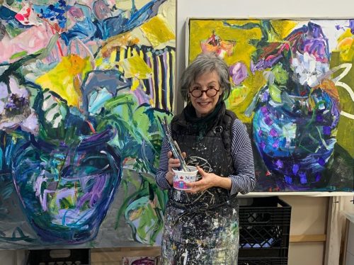 A woman is standing in front of two colourful paintings depicting a vase with blues, greens, purples, and yellows. The woman is holding a jar of paintbrushes and is wearing a paint-covered apron.