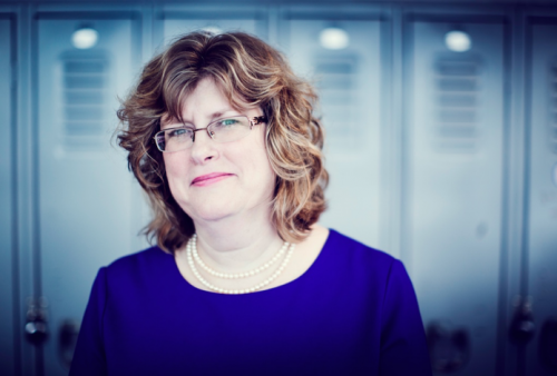 Headhshot of President and CEO, Dr. Ann Marie Vaughan in a blue dress with two pearl necklaces. The background is blurred lockers.