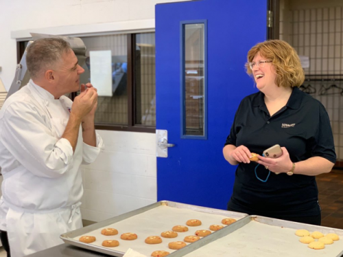 Dr. Ann Marie Vaughan, President and CEO, in a culinary lab, speaking with a professor. A cooking sheet of cookies is on the table between the two.