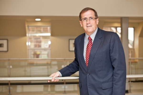 Headshot of Rob Byrne the Senior Vice-President Academic at Loyalist College. He's wearing a suit with a red tie, one hand grabbing the metal bar of a glass railing.