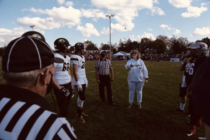 Dr. Ann Marie Vaughan, Loyalist President and CEO, performing a coin flip at a Skyhawks football game. Dr. Vaughan is centre, on either side are football players, and a referee is visible at the left corner of the image.