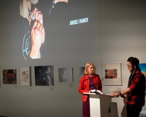 Loyalist College Photojournalism student Andrej Ivanov and Emcee Laura Lynch stand at a podium during a presentation. Lynch is wearing a red jacket, looks down at the podium while Andrej is, wearing a red plaid shirt and scarf, holds a piece of paper. On the wall behind them is a large projected image of a person holding a camera with the name 'Andrej Ivanov' displayed. Framed artwork is visible on the wall below the projection.