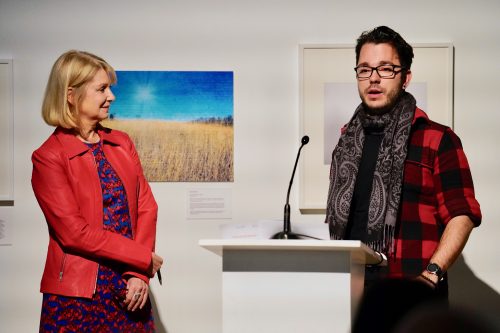 Loyalist College Photojournalism student Andrej Ivanov and Emcee Laura Lynch stand at a podium during a presentation. Lynch is wearing a red jacket and blue dress, smiles while listening to Andrej, who is wearing glasses, a scarf, and a red plaid shirt, as he speaks into the microphone. Framed artwork is displayed on the wall behind them.