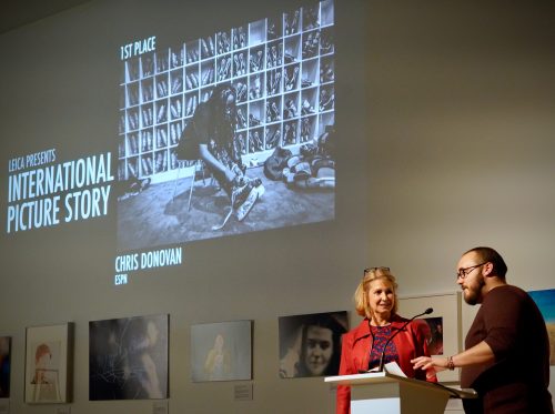 Loyalist College Photojournalism graduate Chris Donovan and Emcee Laura Lynch stand at a podium presenting an award in front of a large projected screen. The screen shows 'LEICA Presents International Picture Story, 1st Place' with a black and white photo of a person sitting in front of shelves. The award is credited to 'Chris Donovan, ESPN.' Several framed photos are displayed on the wall behind them.