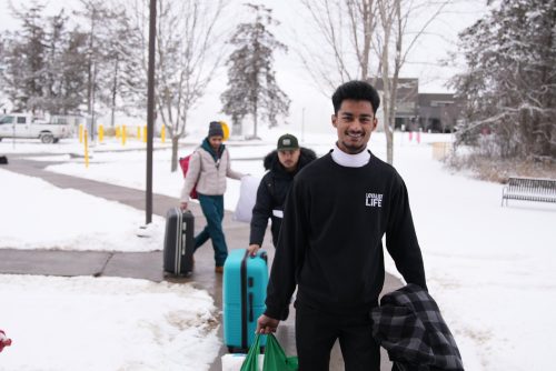 Three students carrying bags and luggage down a sidewalk with snow covering the ground.