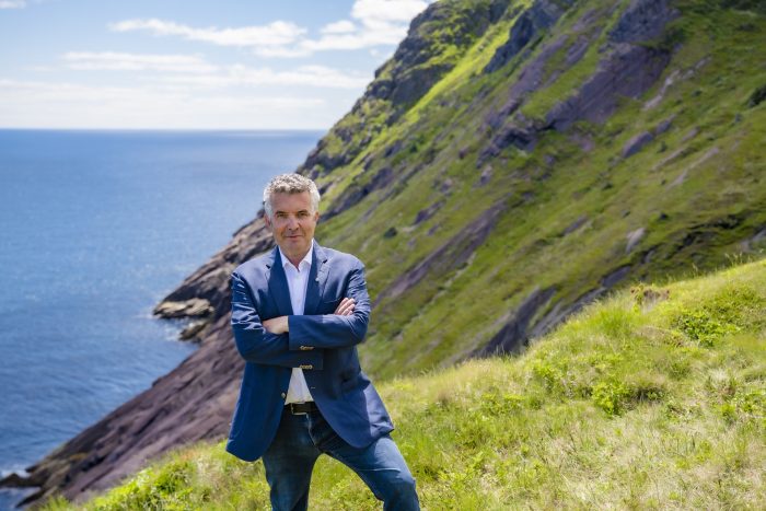 An image of Rick Mercer, standing in a suit with his arms crossed on the side of a hill. In the background is more of the hill going up, and a body of water below.