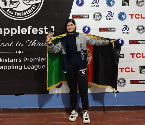 An image of a Loyalist student, Roya Shafai, posing in front of a Grapplefest banner. Roya is holding a flag for Afghanistan.