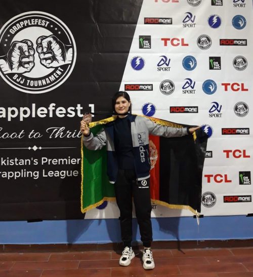 An image of a Loyalist student, Roya Shafai, posing in front of a Grapplefest banner. Roya is holding a flag for Afghanistan.