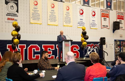 Mark Kirkpatrick on a raised stage at a podium speaking to a large group in the gym at Loyalist College.