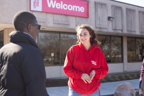 A Loyalist student wearing a red Loyalist hoodie talking to another person in front of the main entrance.