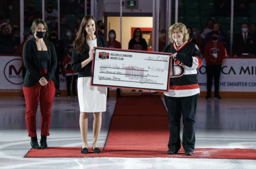 Three women, including Loyalist President and CEO Dr. Ann Marie Vaughan, standing on an ice rink, with two holding a large ceremonial cheque from the Belleville Senators Hockey Club. The cheque is made out to the Loyalist College Foundation for $10,000. The women are dressed in formal attire, and the presentation takes place on a red carpet with spectators in the background.