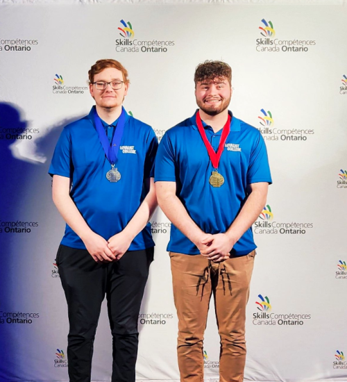 Two Loyalist students posing in front of a Skills Ontario Competition banner. One has a silver medal, the other a gold medal.