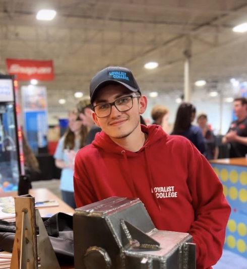 A Loyalist student wearing a red Loyalist hoodie and a black hat leaning against an unknown surface. In the background is a blurred-out warehouse space full of booths and people.