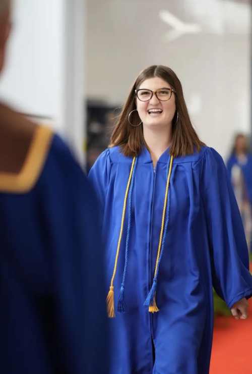 A female student in a blue graduation gown with yellow and blue tassels, walking across the stage smiling.