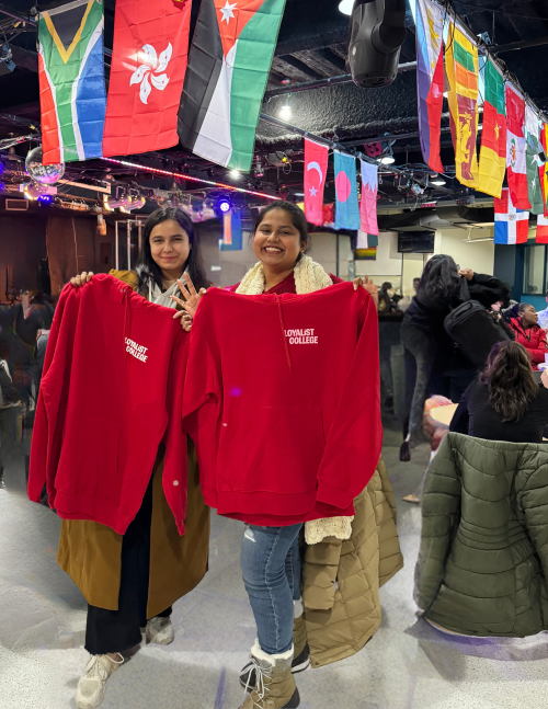 Two students posing with red Loyalist sweaters in the Shark Tank Pub, above them in the background are flags for various countries around the world.