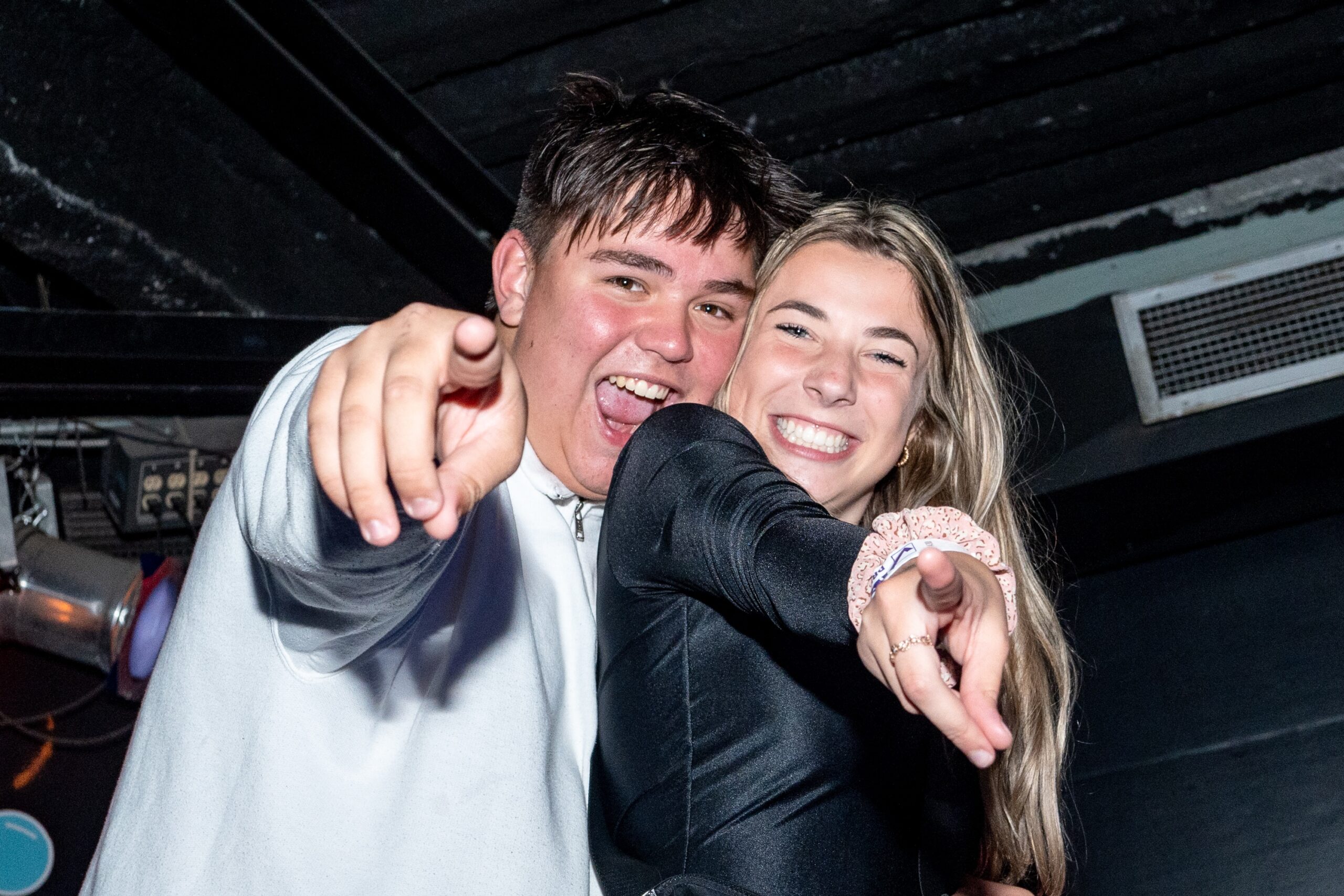 Two smiling individuals point playfully towards the camera during a fun moment at an event. Both appear to be enjoying themselves, with one wearing a white jacket and the other in a black outfit, creating a lively and joyful atmosphere.