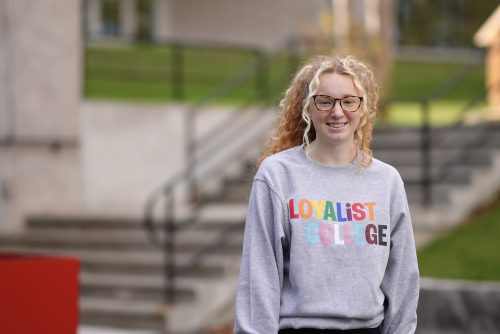 A Loyalist student posing outside by the Shark Tank Patio, wearing a Loyalist College Pride sweatshirt.