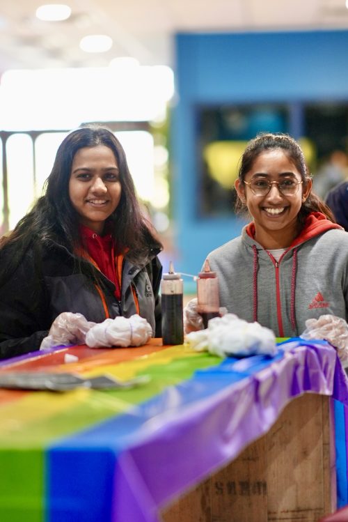 Two students at a tie-dye station with a rainbow tablecloth covering the table, smiling at the camera.