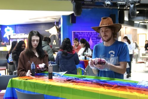 Two students focused on tie-dyeing white clothing with the Shark Tank Pub in the background. Covering the table is a rainbow tablecloth.