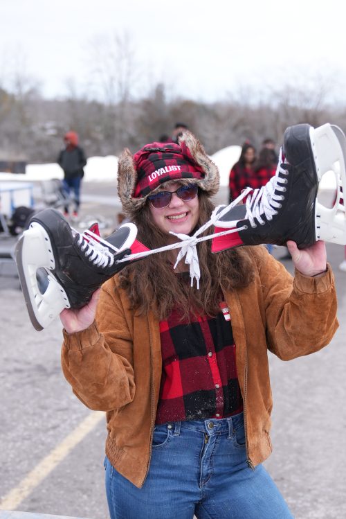 A female student posing with hockey skates in her hands, wearing a red plaid Loyalist hat and shirt.