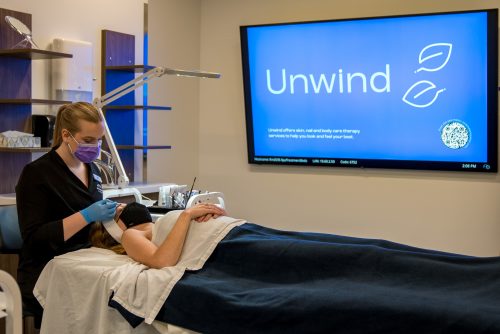 An esthetics student with a patient lying on the table, performing a service. A TV screen in the background shows an image of the Unwind logo and a QR code.