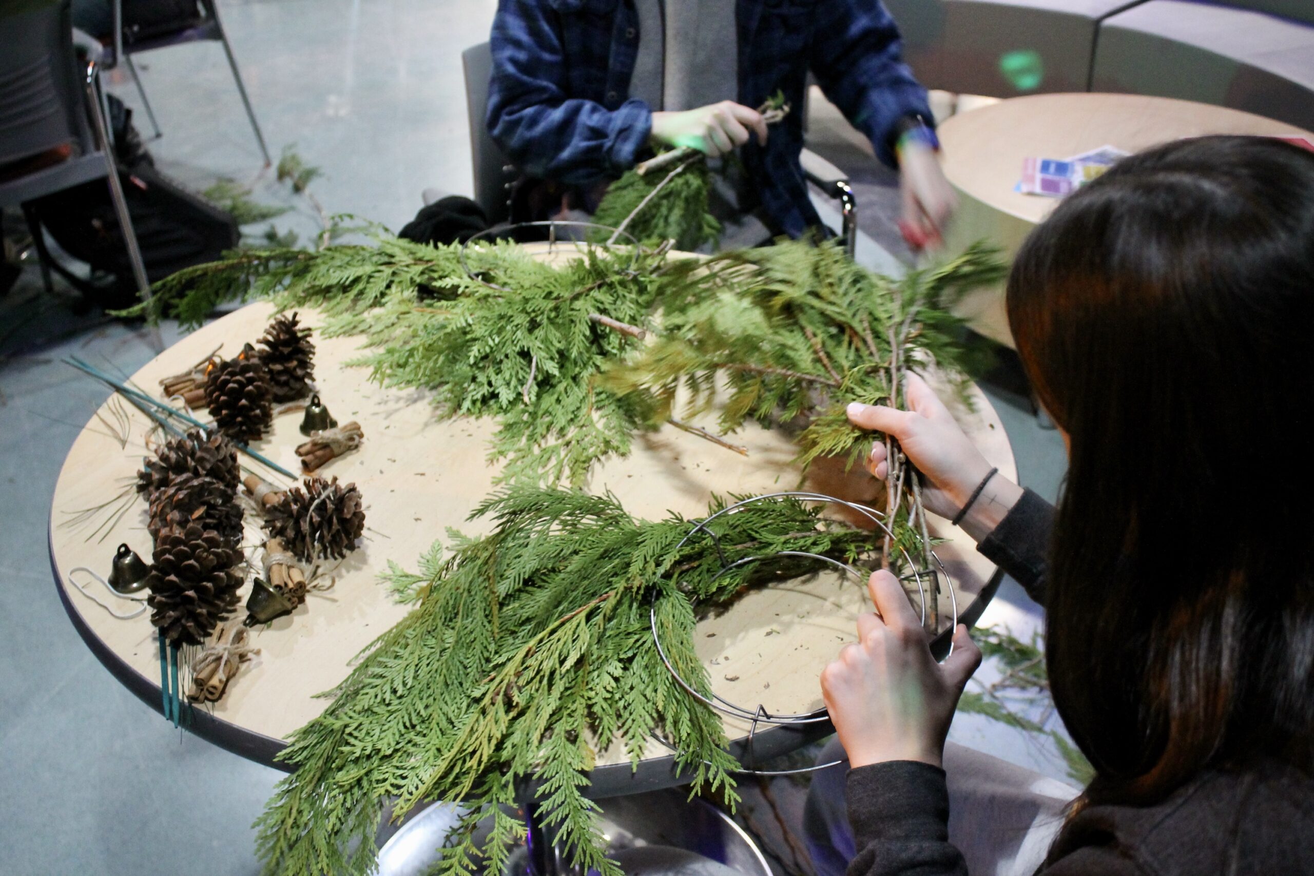 Two students sit at a table assembling wreaths made from fresh branches and pinecones. The table is scattered with natural materials, including twigs, bells, and wire frames for creating the wreaths.