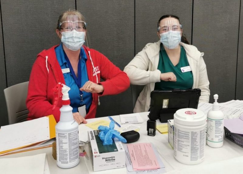 A nursing faculty member and student seated at a table, both wearing face masks, face shields, and ID badges. They are bumping elbows as a greeting. The table is covered with medical supplies including gloves, disinfectant wipes, and paperwork. One worker is wearing a red jacket, while the other is in a green shirt.