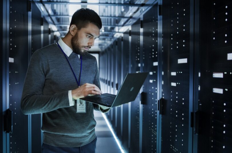 A person works on a laptop in a dimly lit data centre, surrounded by rows of server racks with blinking lights.