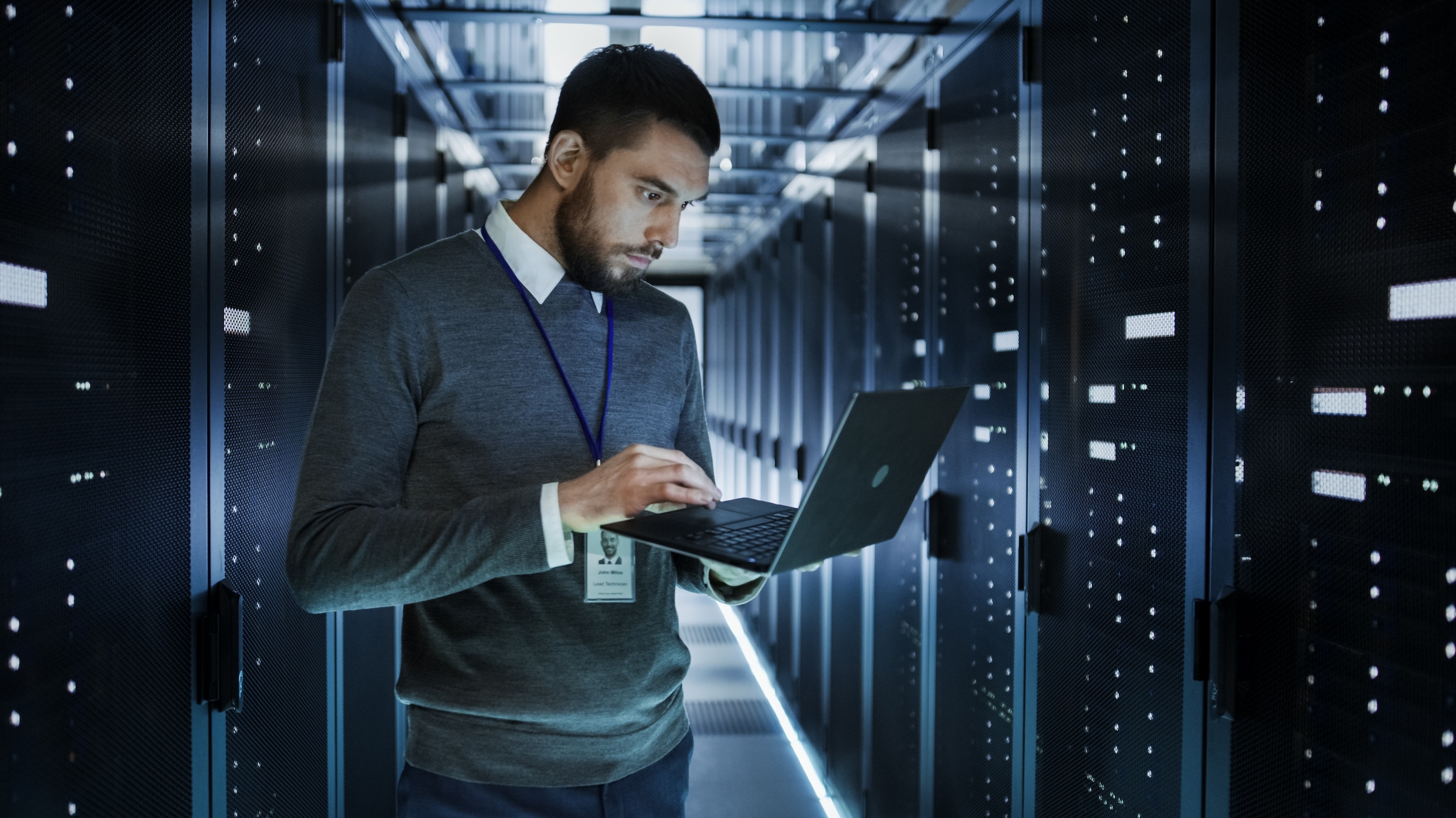 A person works on a laptop in a dimly lit data centre, surrounded by rows of server racks with blinking lights.