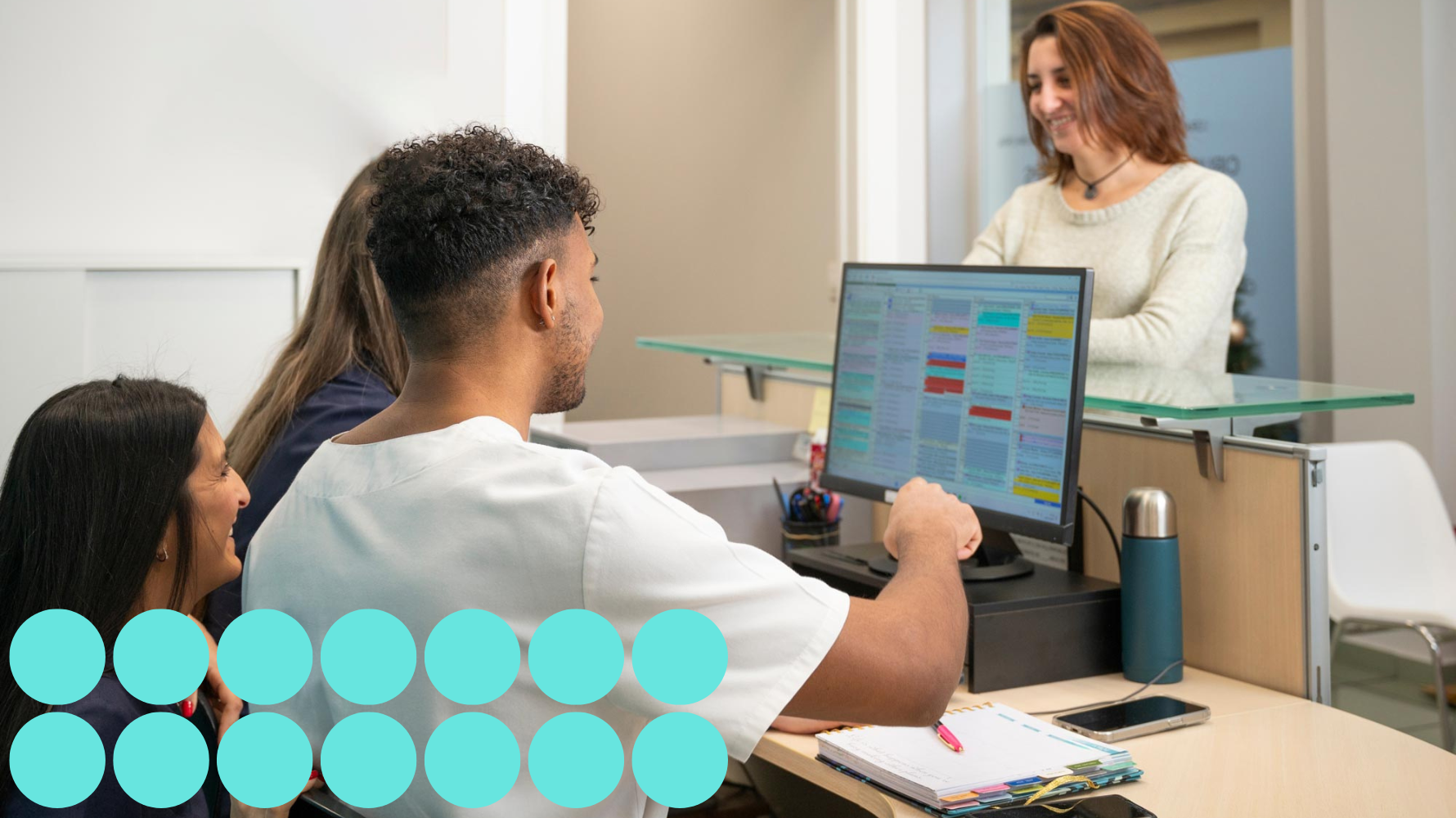 Students learning dental office scheduling and patient interaction at a reception desk. There are 14 small teal circles on the lower left hand side of the image.