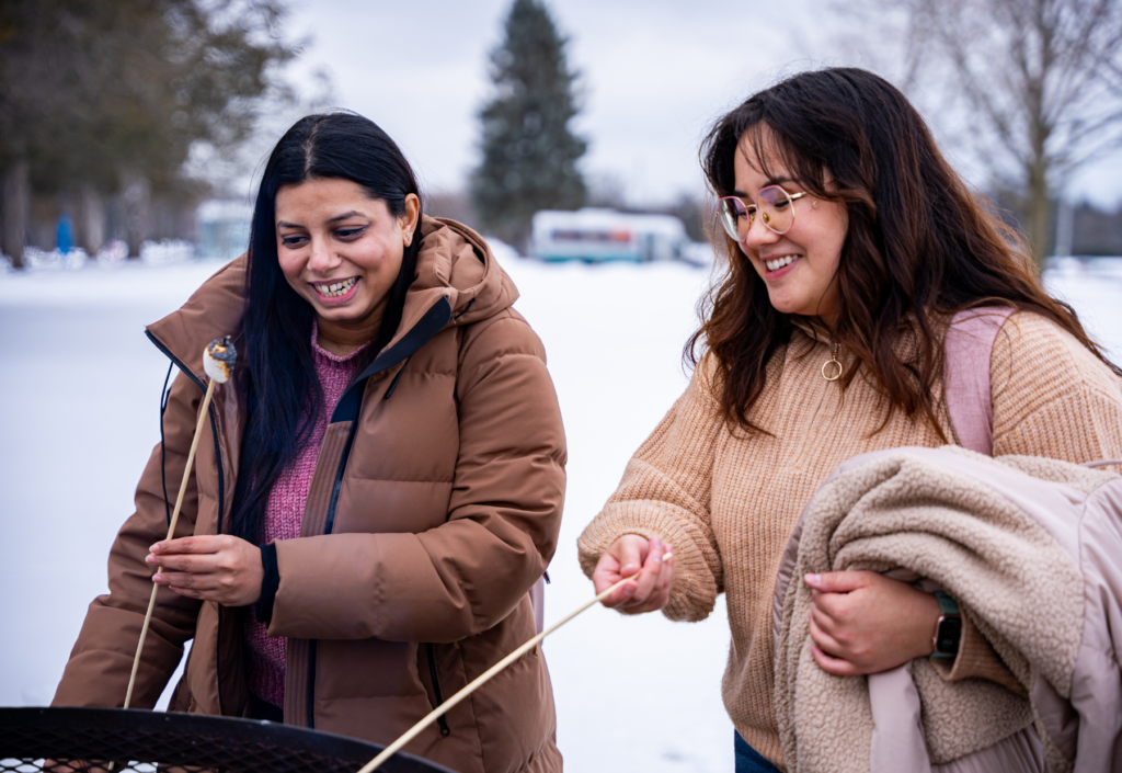 Two students smiling and roasting marshmallows over an outdoor fire pit on a snowy winter day. One is wearing a brown winter coat, while the other is dressed in a beige sweater and carrying a fleece-lined jacket. Behind them is a snow-covered landscape with trees and a parked bus.