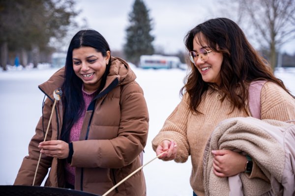 Two students smiling and roasting marshmallows over an outdoor fire pit on a snowy winter day. One is wearing a brown winter coat, while the other is dressed in a beige sweater and carrying a fleece-lined jacket. Behind them is a snow-covered landscape with trees and a parked bus.