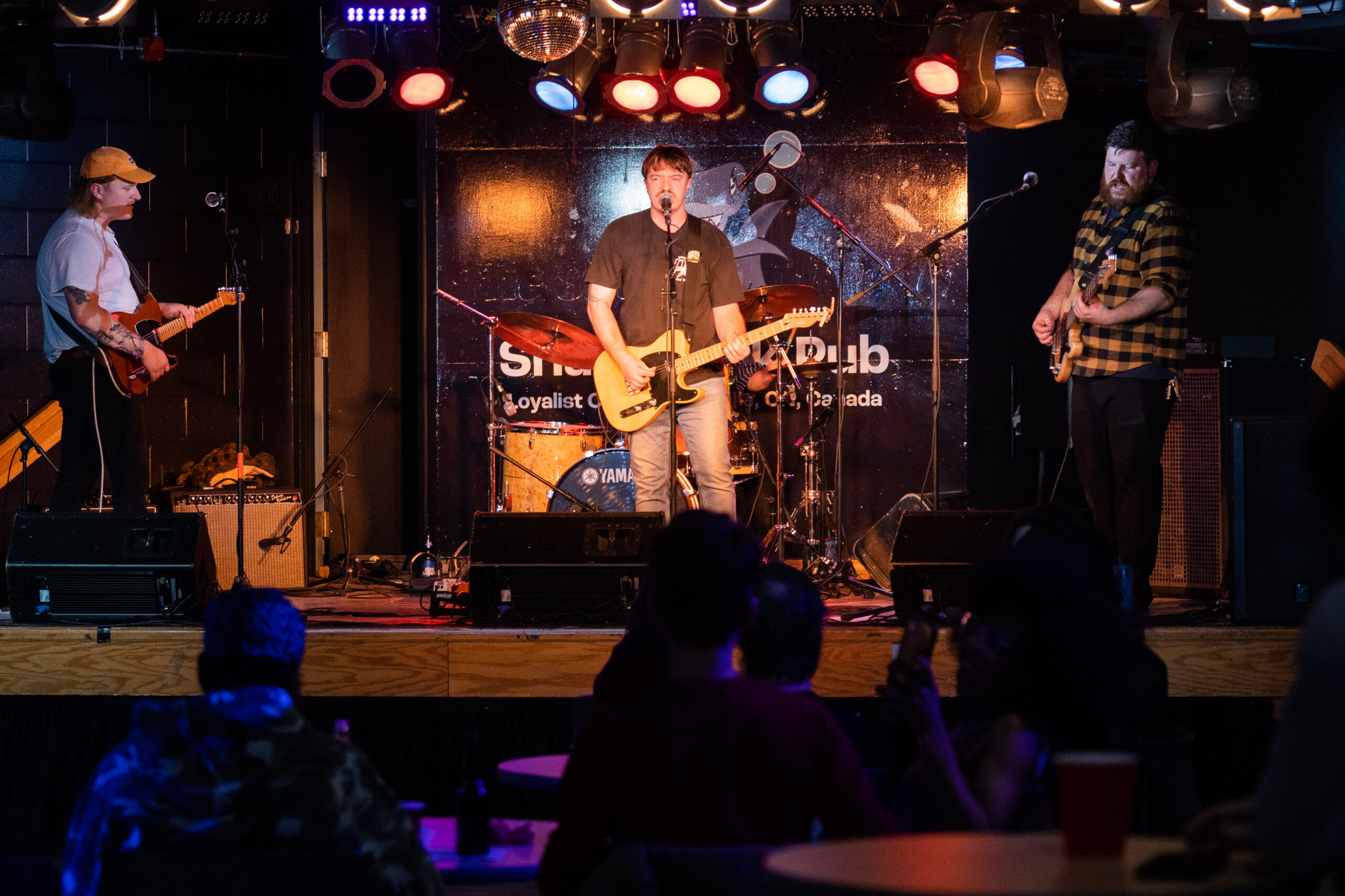 A live band performs on stage at Loyalist College's Shark Tank Pub. The lead singer, standing at the centre, plays an electric guitar while singing into a microphone. He is accompanied by two other musicians, each playing guitars, under colourful stage lights. The background features a drum set and a decorated stage wall with the venue's name. Several audience members are seated at tables, enjoying the performance in the dimly lit venue.