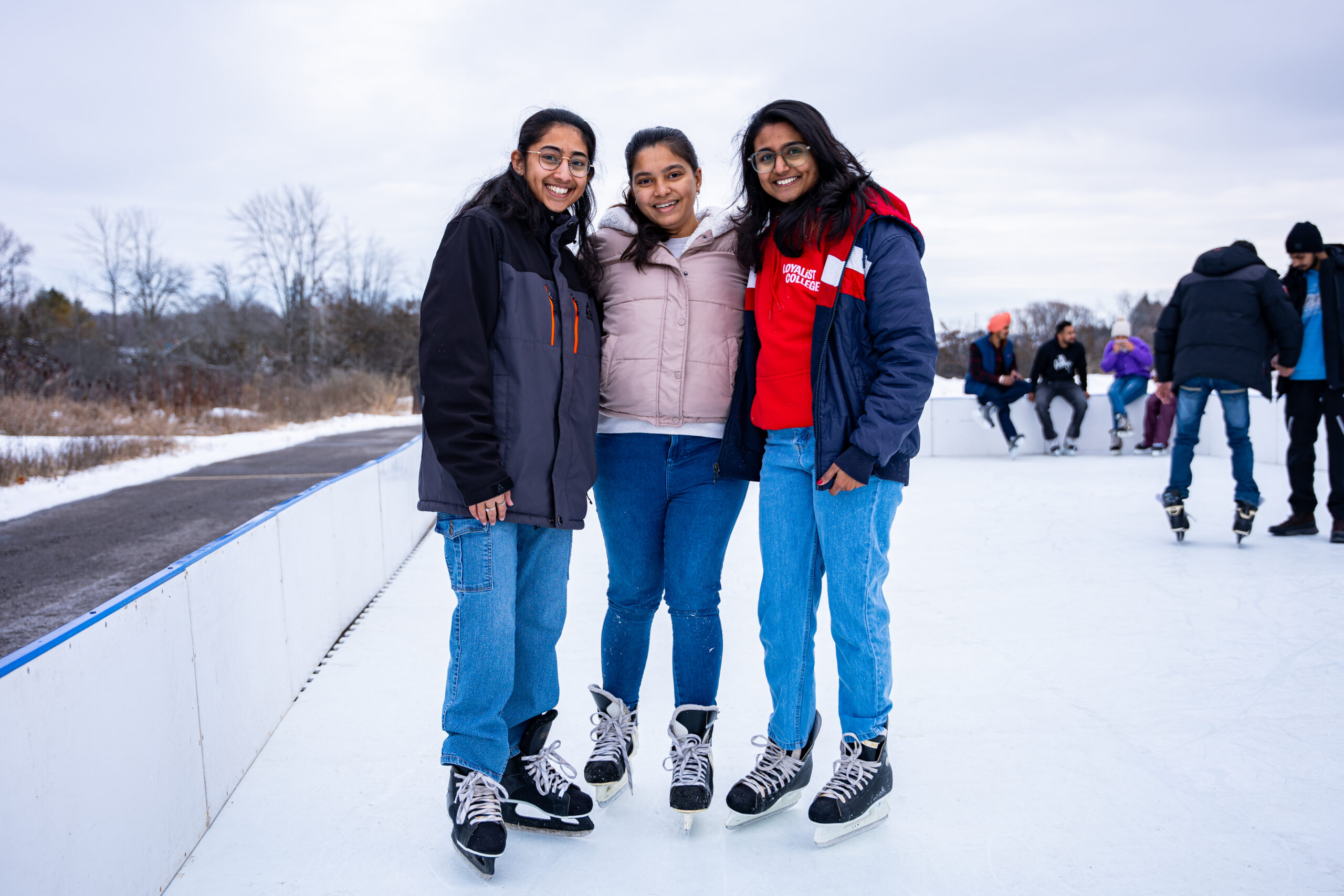 Three young women are smiling and posing together on an outdoor skating rink during the winter season. They are dressed warmly in jackets, jeans, and ice skates. The rink is surrounded by a snowy landscape with bare trees in the background. In the distance, other skaters can be seen enjoying the ice, creating a lively and fun winter scene.