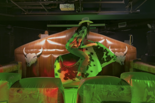 A female student dressed in a green plaid shirt, jeans, and a cowboy hat is riding a mechanical bull at an indoor event. Bright green and red lighting illuminate the scene, adding to the excitement. Spectators can be seen in the background, watching the rider's attempt to stay on the bull.