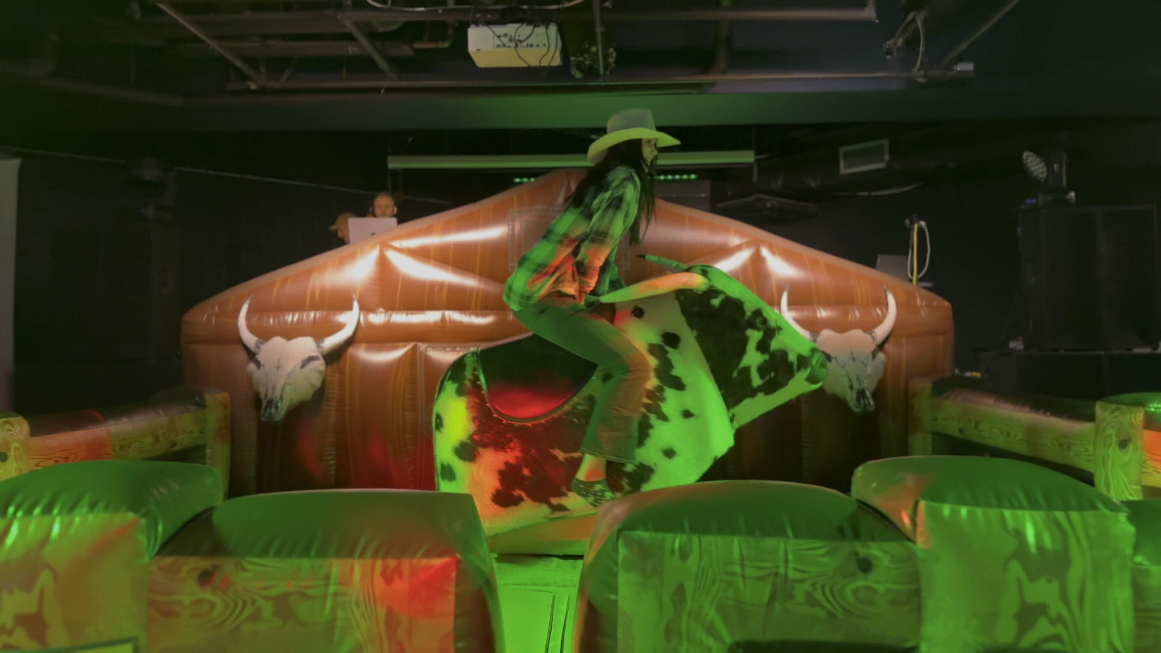 A female student dressed in a green plaid shirt, jeans, and a cowboy hat is riding a mechanical bull at an indoor event. Bright green and red lighting illuminate the scene, adding to the excitement. Spectators can be seen in the background, watching the rider's attempt to stay on the bull.