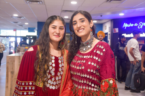 Two women dressed in traditional, vibrant red attire adorned with intricate silver embellishments smile warmly at the camera. Their outfits include detailed embroidery and reflective metallic designs, and they are accessorized with matching jewelry. The event appears to be a celebration of culture and diversity, with other participants in traditional clothing visible in the background of a brightly lit indoor space.