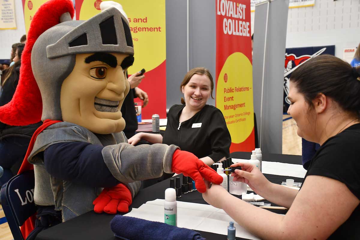 Lanny the Lancer, a mascot, gets a manicure from an Esthetics student at an Open House event. Another student looks on in the background.