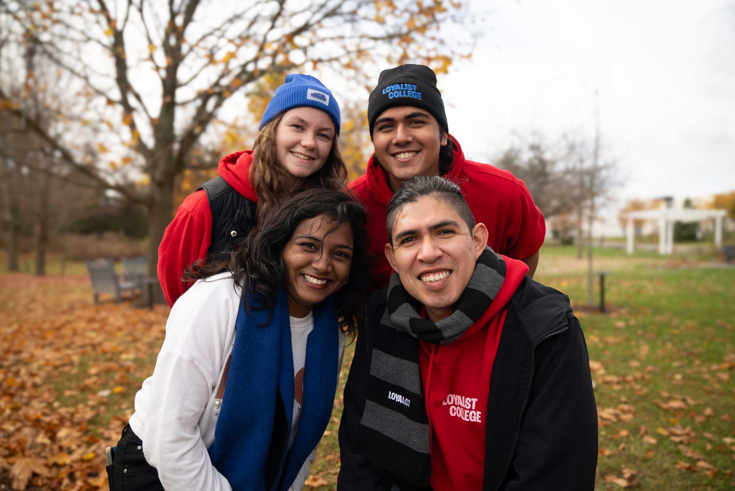 Four Loyalist College students posing together outdoors in a park during autumn, wearing red Loyalist hoodies, scarves, and hats, smiling with fallen leaves in the background.