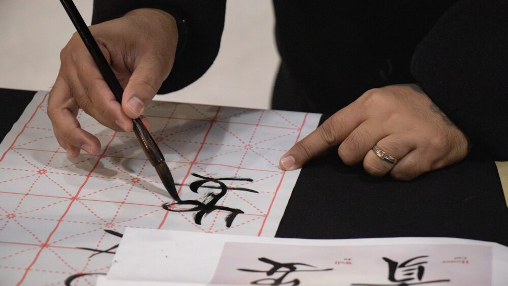 A close-up shot captures hands practicing Chinese calligraphy during a cultural event. One hand holds a brush dipped in black ink, carefully writing Chinese characters on a white practice sheet with a red grid pattern.