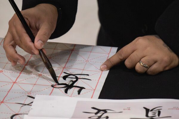 A close-up shot captures hands practicing Chinese calligraphy during a cultural event. One hand holds a brush dipped in black ink, carefully writing Chinese characters on a white practice sheet with a red grid pattern.