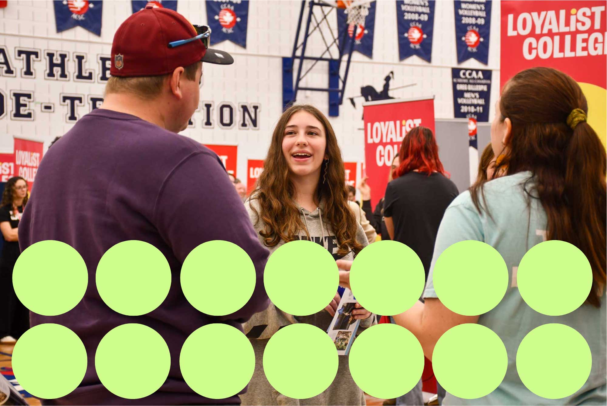 A visitor is engaging with a Loyalist College affiliate. They are holding a pamphlet and in a conversation with the affiliate. There are many Loyalist College signs surrounding the gym.