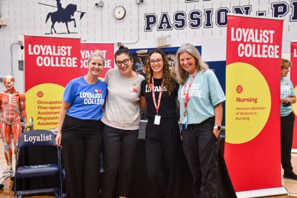 Four Loyalist College Staff members are smiling for a photo. They are standing in front of a table, in the gym. There are many open house banners surrounding them.