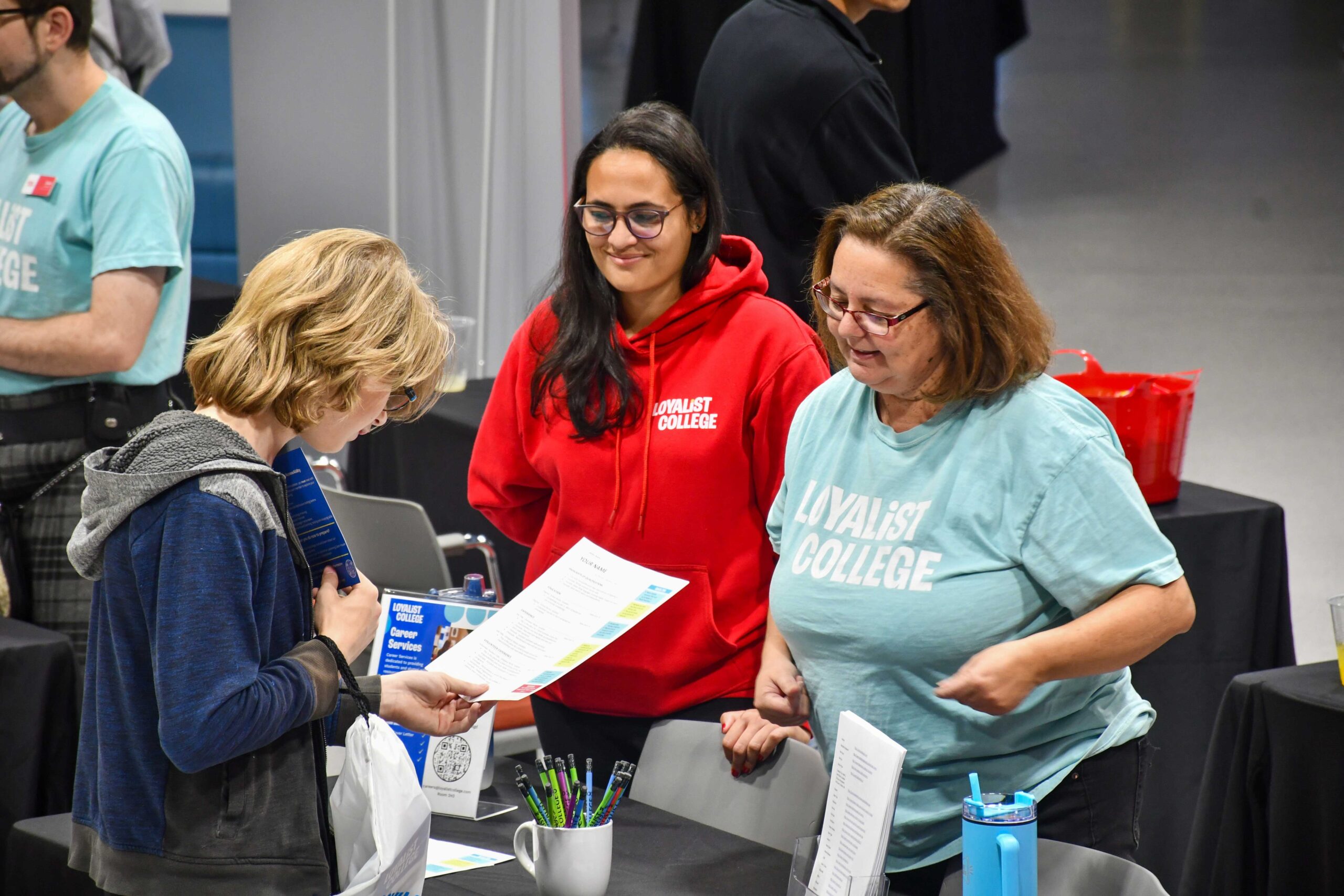 Two Loyalist College Affiliates are standing behind a table at the Open House. A visitor is holding and reading a brochure from their booth and engaging with the affiliates.