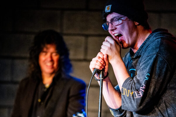 A high-energy moment during a live music performance. The singer is passionately singing into a microphone, wearing a hoodie and beanie, while gripping the mic stand. Behind him, a guitarist smiles while playing an electric guitar. The background is dimly lit and the focus on the musicians.